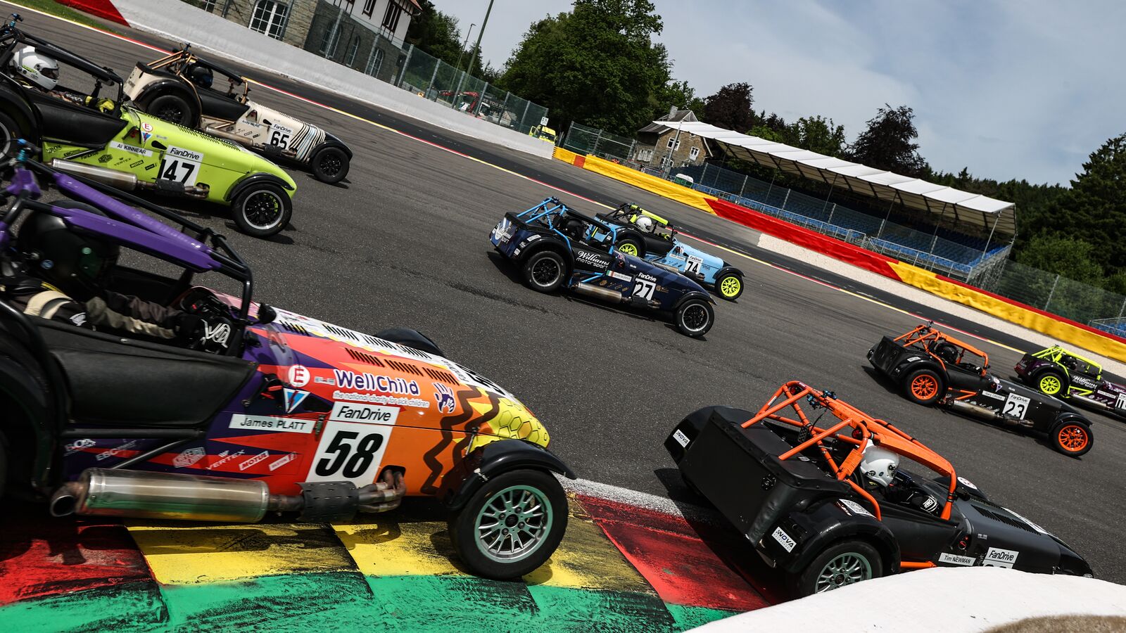 A pack of Caterham Seven race cars bunched through a hairpin, led by the orange and purple #58 on the kerb